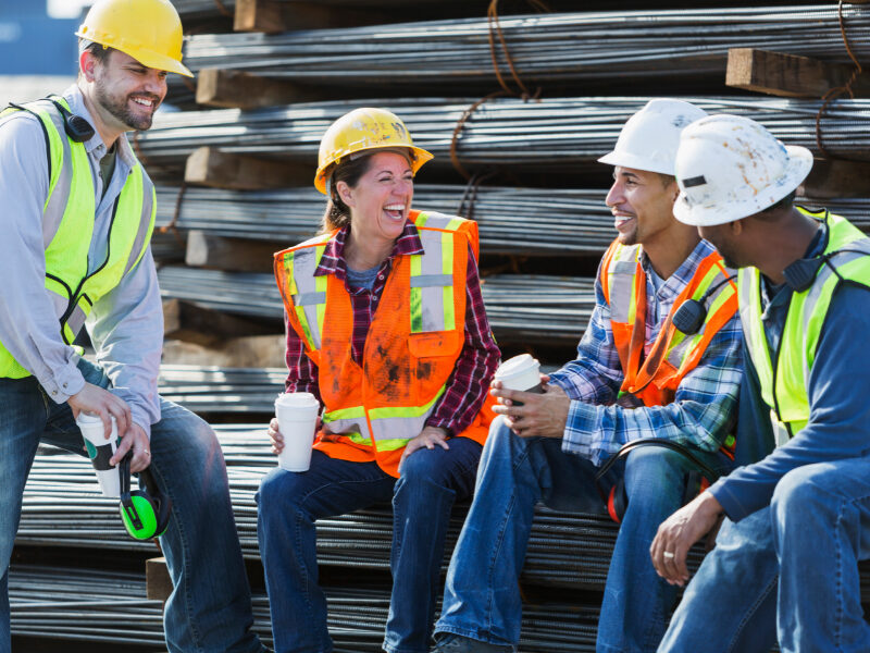 A multi-ethnic group of workers, a woman and three men, wearing safety vests and hardhats taking a coffee break. They are sitting on a stack of steel rebars (construction material), drinking coffee and talking.