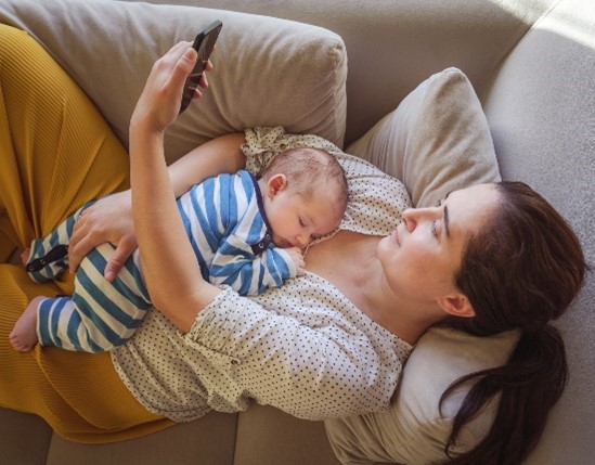Mother looking at her phone with baby, laying down on couch.